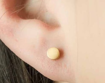 May include: A small, circular, gold-colored stud earring with a smooth, flat surface. The earring is positioned in the lower lobe of an ear, featuring a simple, minimalist design that sits flush against the skin with a polished, metallic finish.