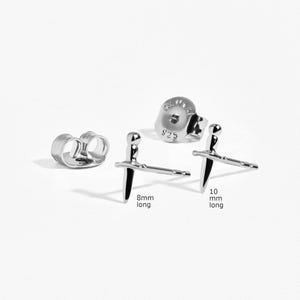 May include: Two silver-toned sword-shaped stud earrings with matching earring backs. One earring post is labeled 8mm long and the other is labeled 10mm long. The earring backs are stamped with 925, indicating sterling silver composition.
