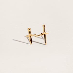 May include: A pair of gold-toned stud earrings shaped like miniature daggers. Each earring features a rounded pommel, a horizontal crossguard, and a pointed blade. The earrings have a polished, reflective metallic finish and include thin posts.