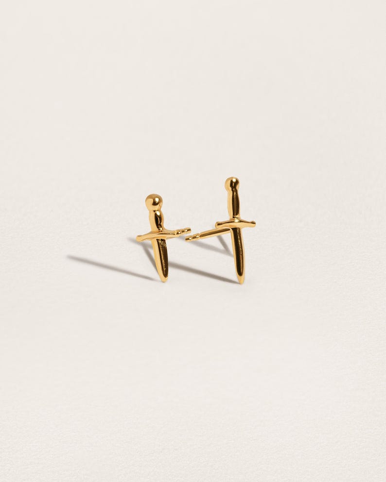 May include: A pair of gold-toned stud earrings shaped like miniature daggers. Each earring features a rounded pommel, a horizontal crossguard, and a pointed blade. The earrings have a polished, reflective metallic finish and include thin posts.