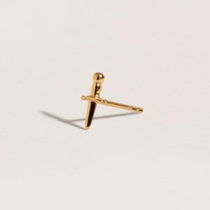 May include: A gold stud earring shaped like a small dagger or sword. The earring features a rounded pommel, a horizontal crossguard, and a pointed blade. A thin post extends from the back of the crossguard, designed for insertion into an earlobe.