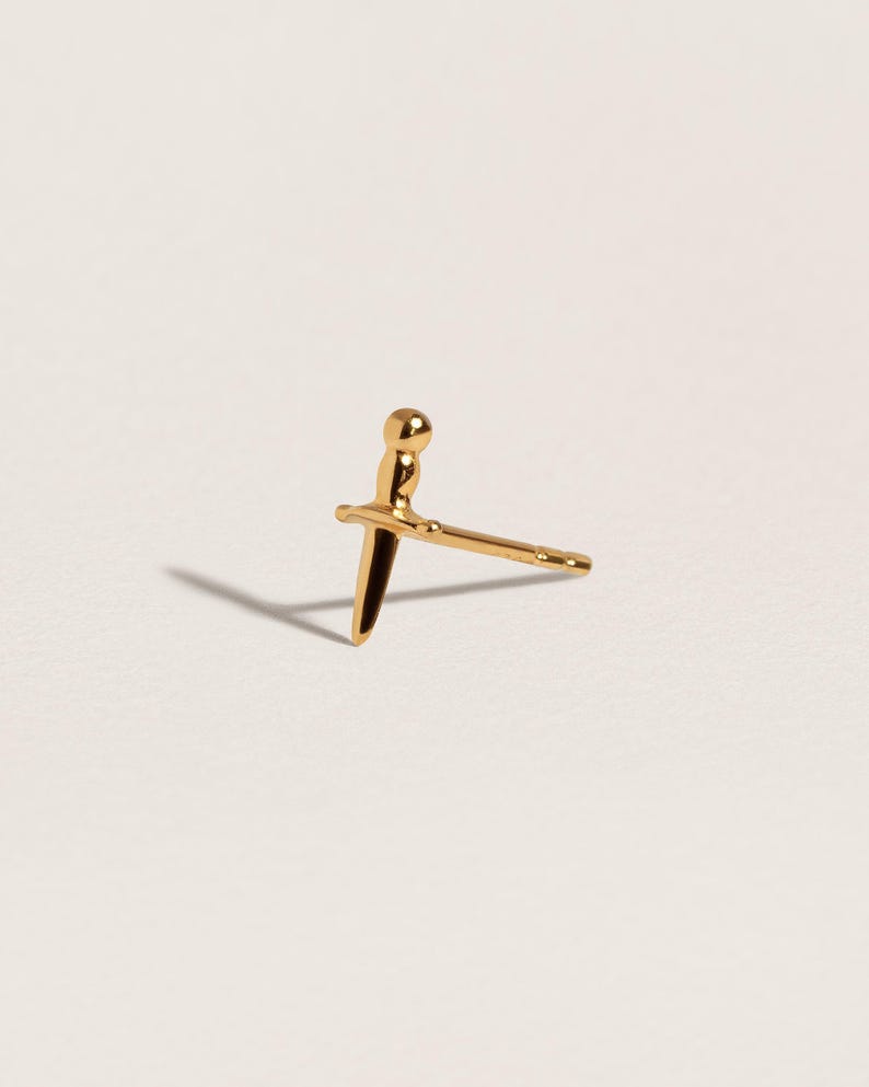 May include: A gold stud earring shaped like a small dagger or sword. The earring features a rounded pommel, a horizontal crossguard, and a pointed blade. A thin post extends from the back of the crossguard, designed for insertion into an earlobe.