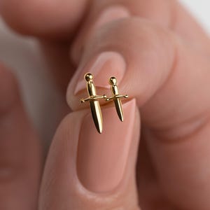 May include: Two gold-toned stud earrings shaped like miniature daggers. The earrings feature a polished metallic finish with a rounded pommel, a crossguard, and a pointed blade. The two daggers are of slightly different sizes, both gold in color.