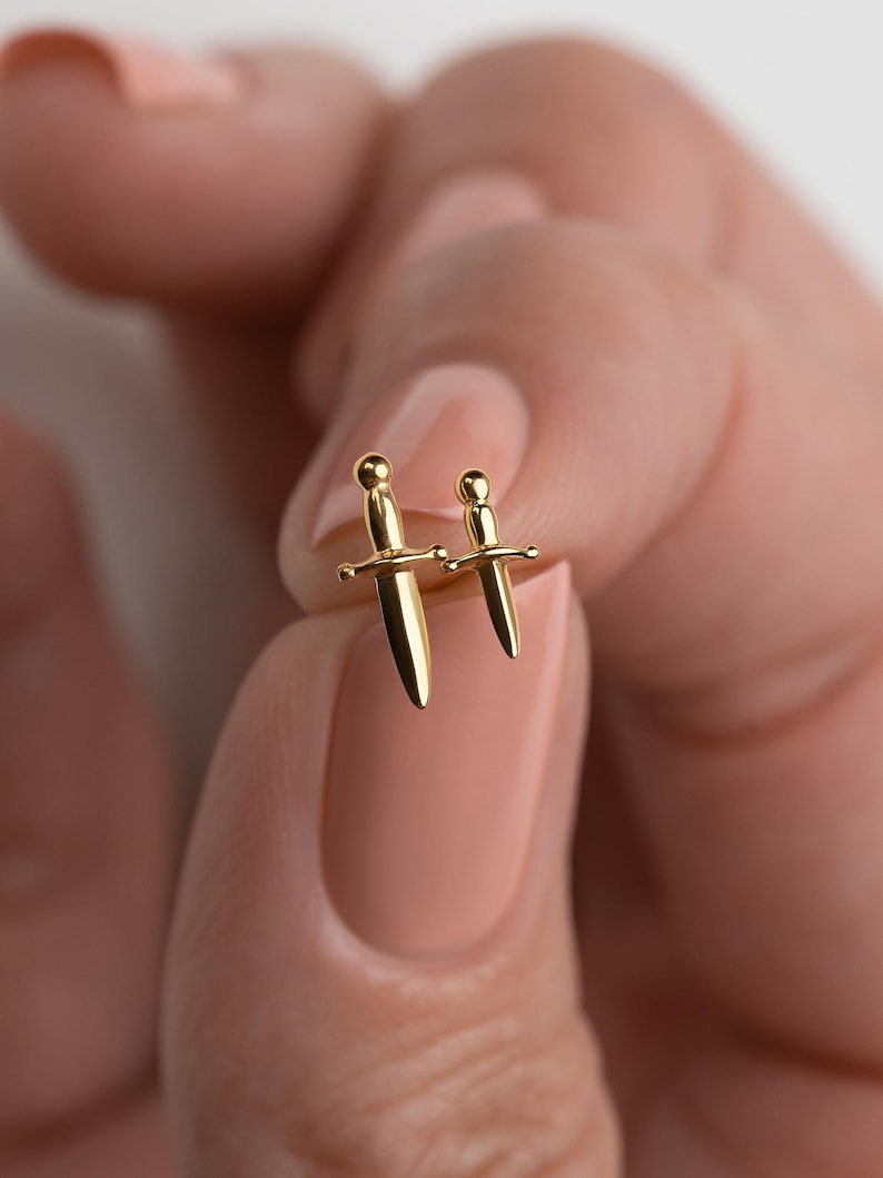 May include: Two gold-toned stud earrings shaped like miniature daggers. The earrings feature a polished metallic finish with a rounded pommel, a crossguard, and a pointed blade. The two daggers are of slightly different sizes, both gold in color.