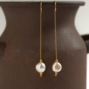 May include: A pair of gold chain earrings with white pearl drops. The earrings are hanging from a brown ceramic vase.