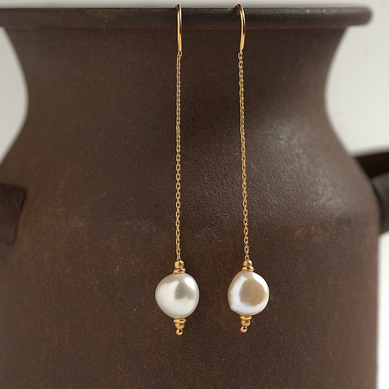 May include: A pair of gold chain earrings with white pearl drops. The earrings are hanging from a brown ceramic vase.
