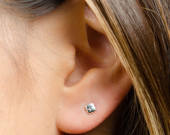 May include: A small, polished silver-toned square stud earring with rounded corners, securely fastened in the earlobe. The earring features a smooth, reflective metallic surface that catches the light, presenting a simple and clean geometric design.
