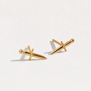 May include: A pair of gold-toned stud earrings shaped like miniature daggers. Each earring features a pointed blade, a crossguard, and a rounded pommel, with a post backing extending from the center of the hilt for secure attachment to the earlobe.