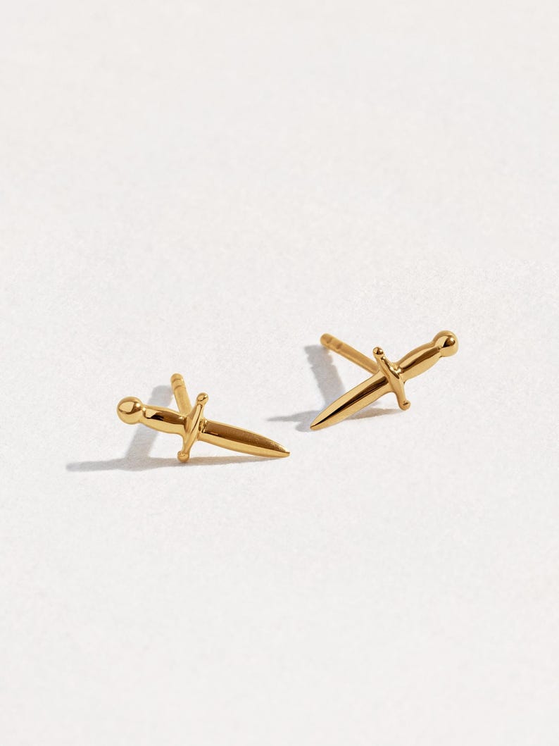 May include: A pair of gold-toned stud earrings shaped like miniature daggers. Each earring features a pointed blade, a crossguard, and a rounded pommel, with a post backing extending from the center of the hilt for secure attachment to the earlobe.
