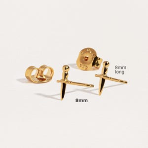 May include: A pair of gold-toned stud earrings shaped like small swords, each measuring 8mm long. The set includes two matching earring backs, one of which is clearly visible with the text 925 stamped on its surface, indicating sterling silver base.