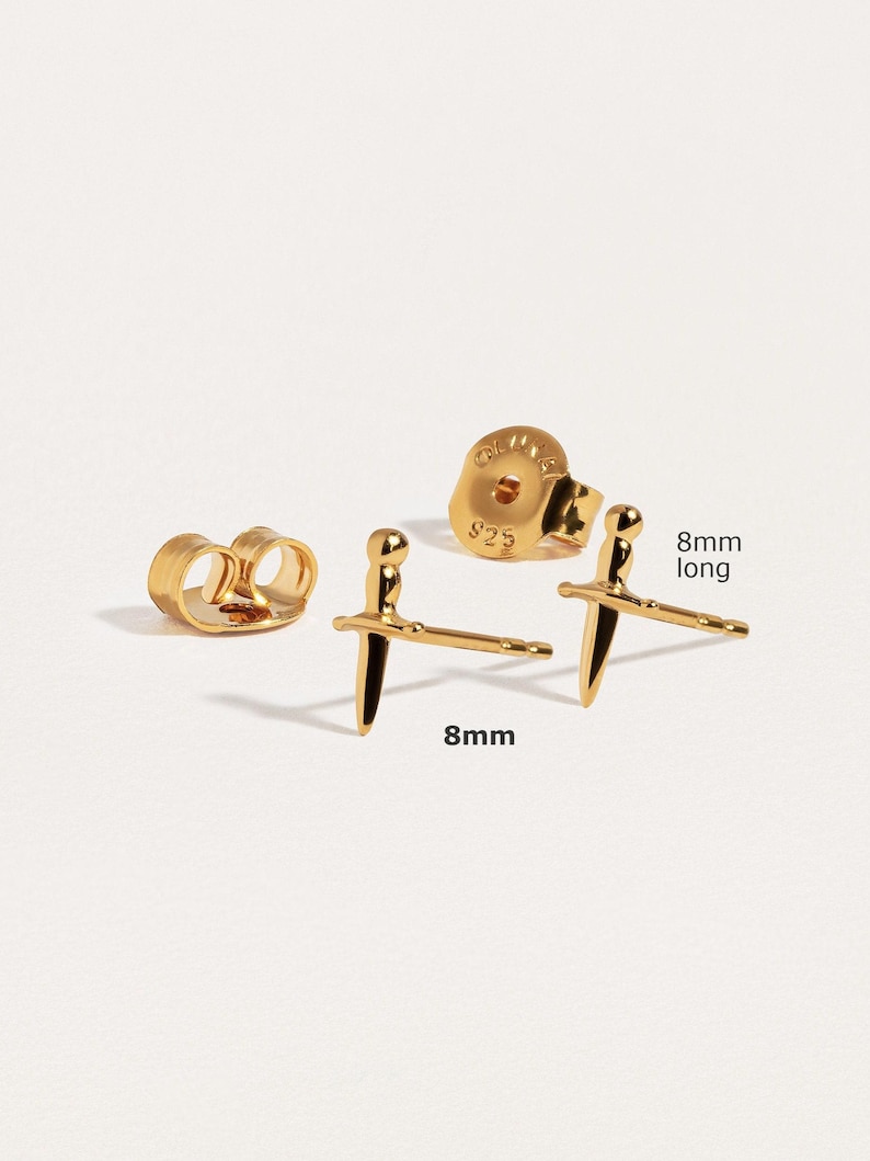 May include: A pair of gold-toned stud earrings shaped like small swords, each measuring 8mm long. The set includes two matching earring backs, one of which is clearly visible with the text 925 stamped on its surface, indicating sterling silver base.