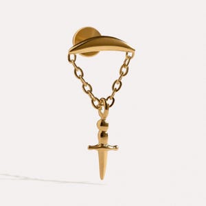 May include: A gold-toned earring featuring a curved bar stud connected by a delicate chain to a small, pointed dagger-shaped pendant. The piece has a polished metallic finish and a secure flat backing, showcasing a clean, structured design.