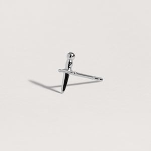 May include: A silver-toned stud earring shaped like a small dagger or sword. The earring features a rounded pommel, a horizontal crossguard, and a pointed blade that extends into a straight post for insertion, with a polished metallic finish.