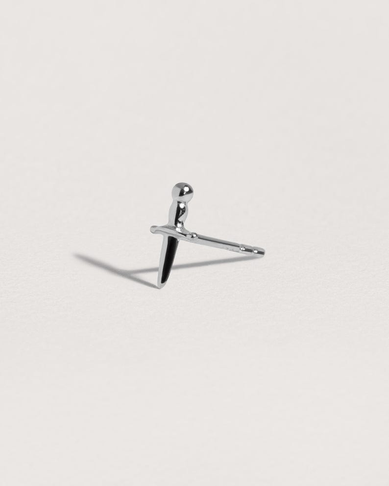 May include: A silver-toned stud earring shaped like a small dagger or sword. The earring features a rounded pommel, a horizontal crossguard, and a pointed blade that extends into a straight post for insertion, with a polished metallic finish.