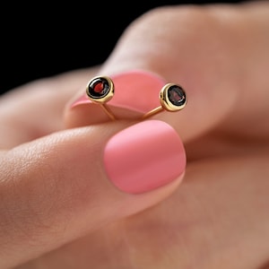 Pode incluir: Um par de brincos de tarracha com pedras preciosas granadas redondas de cor vermelho-escuro, engastadas em aros de metal dourado polido. Cada brinco tem um pino fino e reto que se estende da parte de trás do engaste circular.
