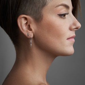 Könnte beinhalten: Eine Frau mit kurzen, dunklen Haaren und einer rasierten Seite, die einen silbernen Dolch-Ohrring trägt. Der Ohrring ist ein Ring mit einem kleinen Dolch-Anhänger.