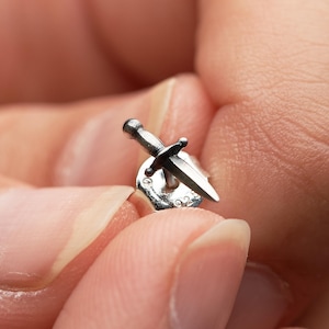 May include: A silver stud earring shaped like a small dagger with a pointed blade and a crossguard. The earring features a metallic finish and is attached to a standard butterfly earring back, which is clearly stamped with the number 925.