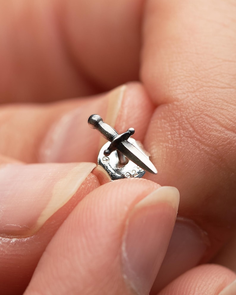 May include: A silver stud earring shaped like a small dagger with a pointed blade and a crossguard. The earring features a metallic finish and is attached to a standard butterfly earring back, which is clearly stamped with the number 925.