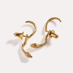 May include: A pair of gold-toned earrings shaped like snakes with textured scales. Each earring features a curved body that mimics a slithering snake, complete with a defined head and a post back with a matching gold-toned butterfly clutch closure.