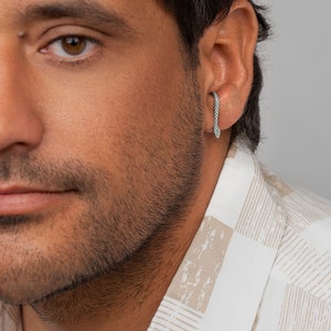 Può includere: Un ear cuff color argento a forma di serpente sottile con una superficie testurizzata simile a scaglie. L'accessorio metallico avvolge il bordo esterno della cartilagine dell'orecchio, presentando una testa appuntita e un corpo ricurvo.