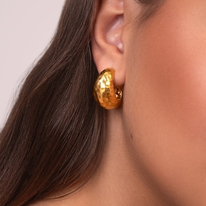 May include: Close-up of a gold-coloured, hammered-texture hoop earring. The earring is a half-circle shape and has a textured surface. The earring is worn on the ear of a person with brown hair.