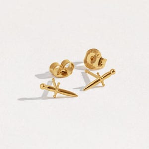 May include: A pair of gold-toned stud earrings shaped like small daggers or swords. Each earring features a pointed blade, a crossguard, and a rounded pommel. Two matching gold-toned earring backs are included, with one showing the stamp 925.