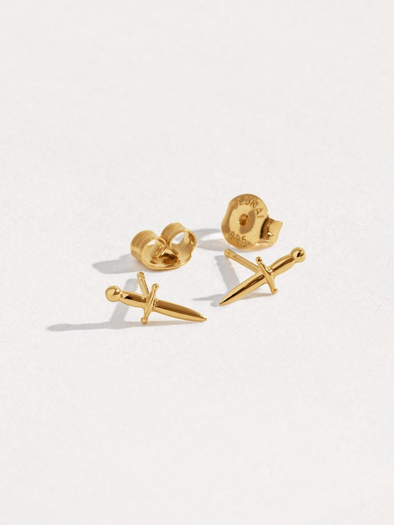 May include: A pair of gold-toned stud earrings shaped like small daggers or swords. Each earring features a pointed blade, a crossguard, and a rounded pommel. Two matching gold-toned earring backs are included, with one showing the stamp 925.