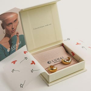 May include: A pair of gold-toned hoop earrings resting inside an open cream-colored jewelry box. The box features a pink fabric insert with the text LUNAI JEWELRY and the phrase A shared dream, made real. The packaging includes white paper with red floral patterns.