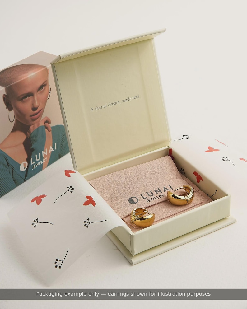 May include: A pair of gold-toned hoop earrings resting inside an open cream-colored jewelry box. The box features a pink fabric insert with the text LUNAI JEWELRY and the phrase A shared dream, made real. The packaging includes white paper with red floral patterns.