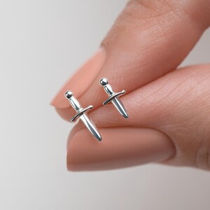 May include: A pair of small, polished silver stud earrings shaped like miniature daggers. Each earring features a rounded pommel, a horizontal crossguard, and a pointed blade, reflecting light with a smooth, metallic finish on the surface.