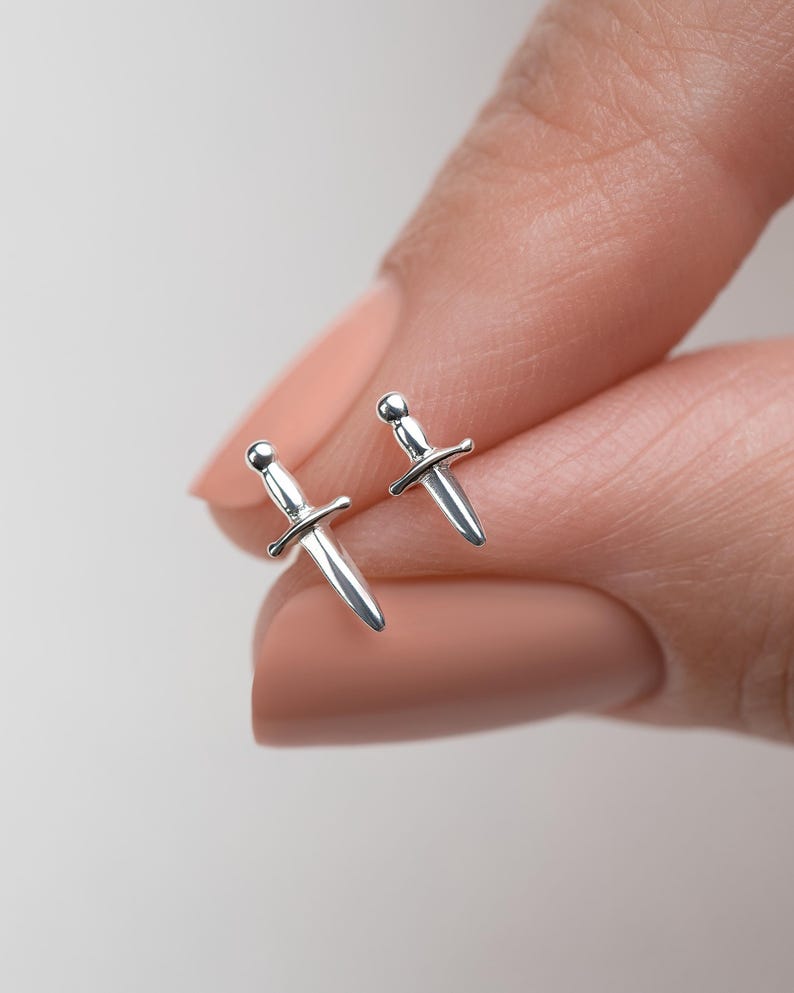 May include: A pair of small, polished silver stud earrings shaped like miniature daggers. Each earring features a rounded pommel, a horizontal crossguard, and a pointed blade, reflecting light with a smooth, metallic finish on the surface.