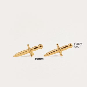 May include: A pair of gold-colored stud earrings shaped like small daggers. Each earring features a pointed blade, a crossguard, and a rounded pommel. The text 10mm long is printed next to the earrings, indicating a length of 10 millimeters.