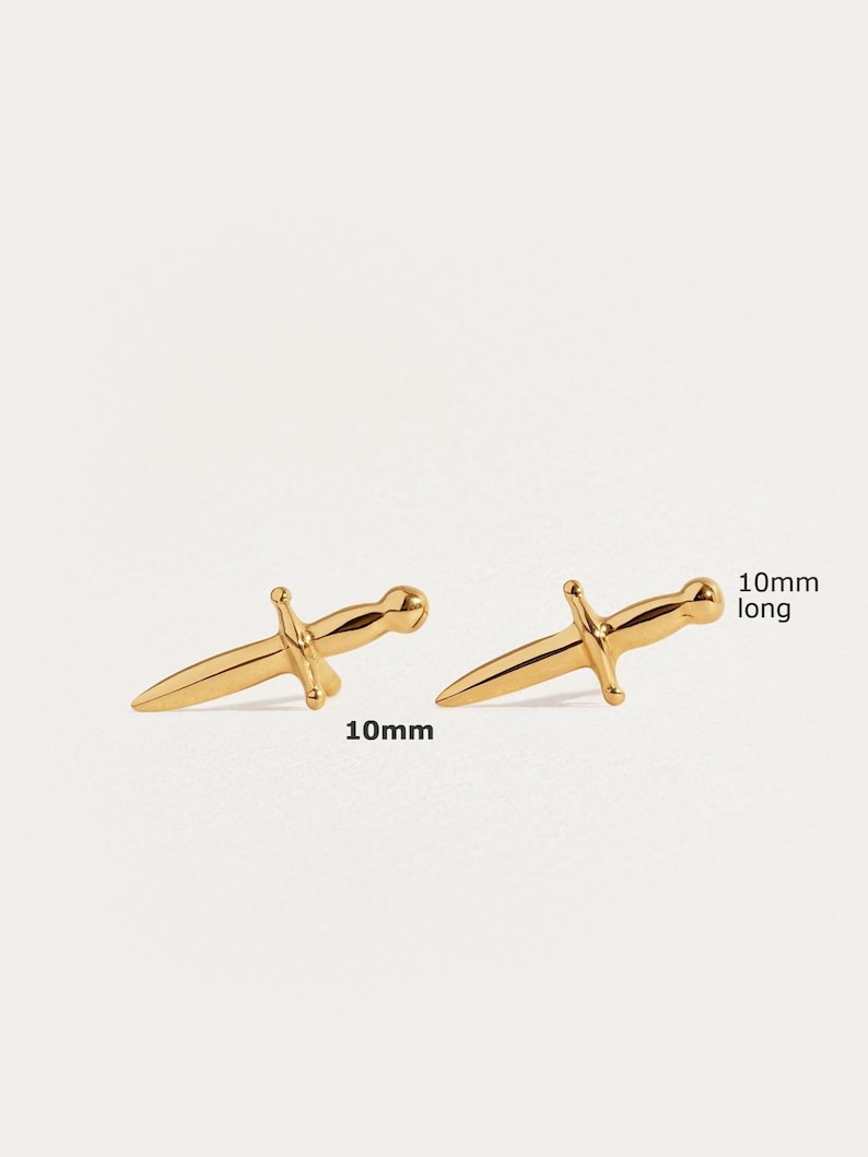 May include: A pair of gold-colored stud earrings shaped like small daggers. Each earring features a pointed blade, a crossguard, and a rounded pommel. The text 10mm long is printed next to the earrings, indicating a length of 10 millimeters.