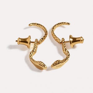 May include: A pair of gold-toned snake-shaped earrings featuring a textured scale pattern and a curved body. Each earring includes a post back closure and the word LUNA is engraved on the underside of the snake head, visible on the right earring.