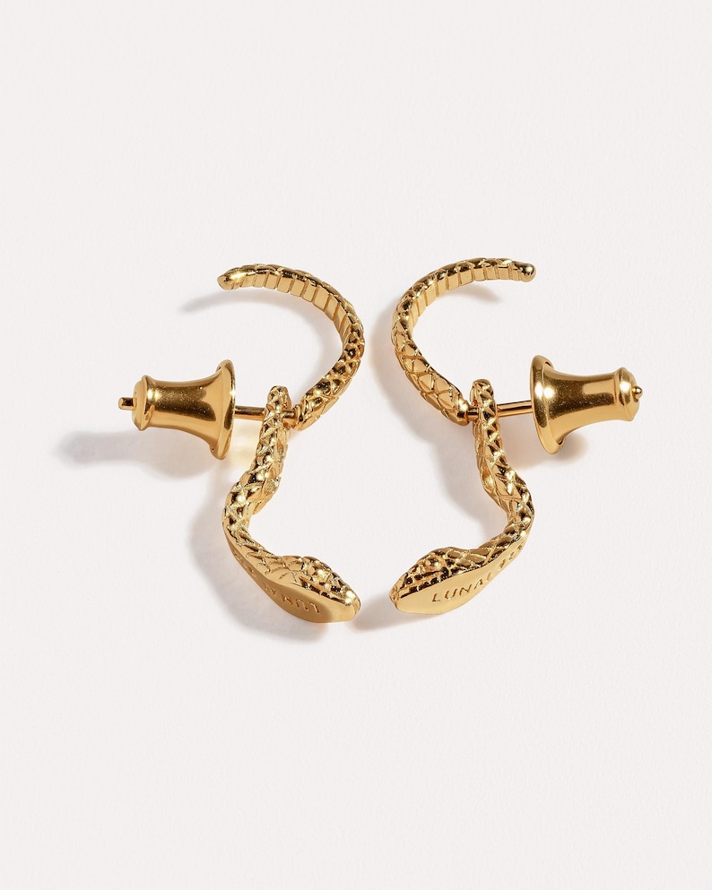 May include: A pair of gold-toned snake-shaped earrings featuring a textured scale pattern and a curved body. Each earring includes a post back closure and the word LUNA is engraved on the underside of the snake head, visible on the right earring.