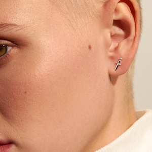 May include: A small silver stud earring shaped like a miniature dagger or sword. The earring features a pointed blade, a central crossguard, and a small handle, all rendered in a polished metallic silver finish, worn in the earlobe.