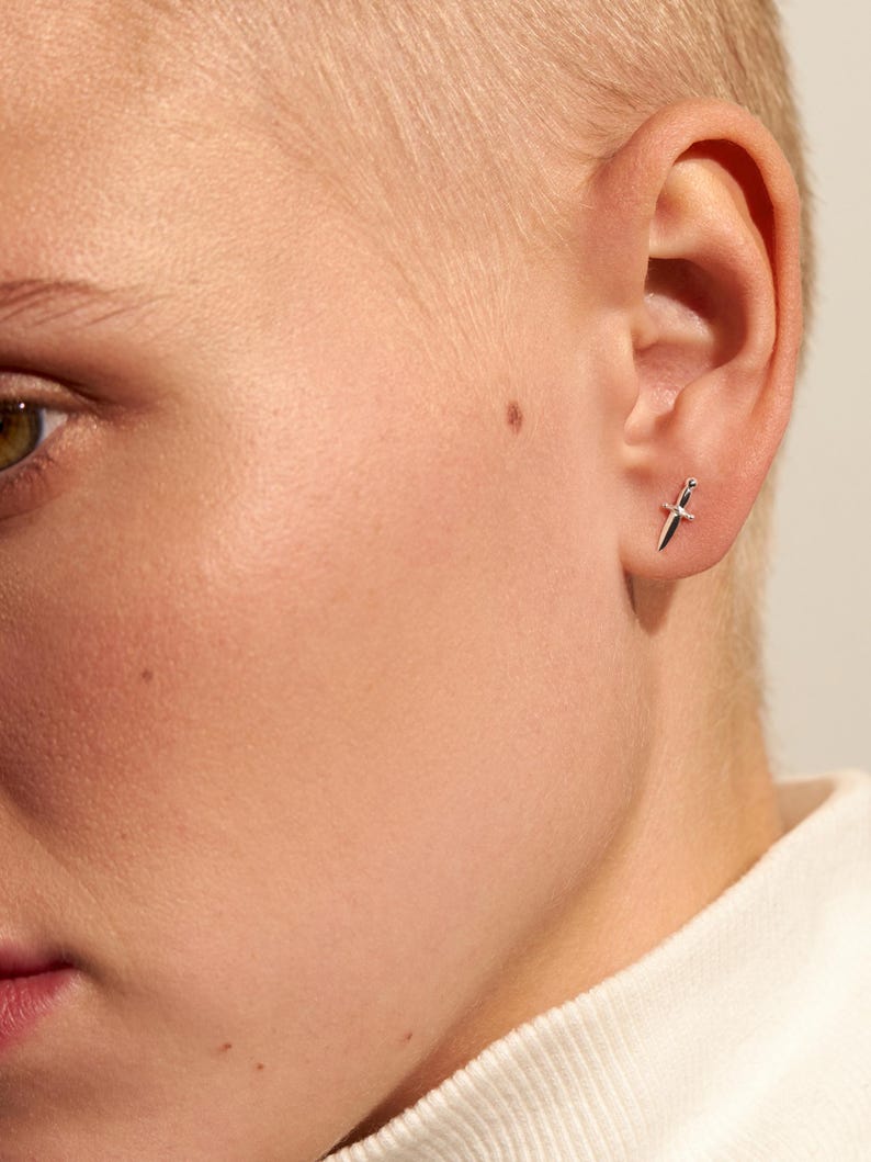 May include: A small silver stud earring shaped like a miniature dagger or sword. The earring features a pointed blade, a central crossguard, and a small handle, all rendered in a polished metallic silver finish, worn in the earlobe.