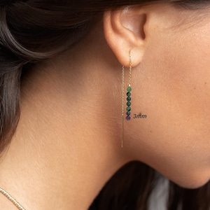 May include: A gold threader earring featuring a vertical row of five small, round, multi-colored beads in shades of green and purple. The beads measure 3mm in diameter. The earring consists of a thin gold chain that passes through the earlobe.