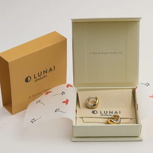 May include: A pair of two-tone gold and silver hoop earrings resting inside an open cream-colored jewelry box. The box lid features the text A shared dream, made real. The box includes a branded insert with the text LUNAI JEWELRY.