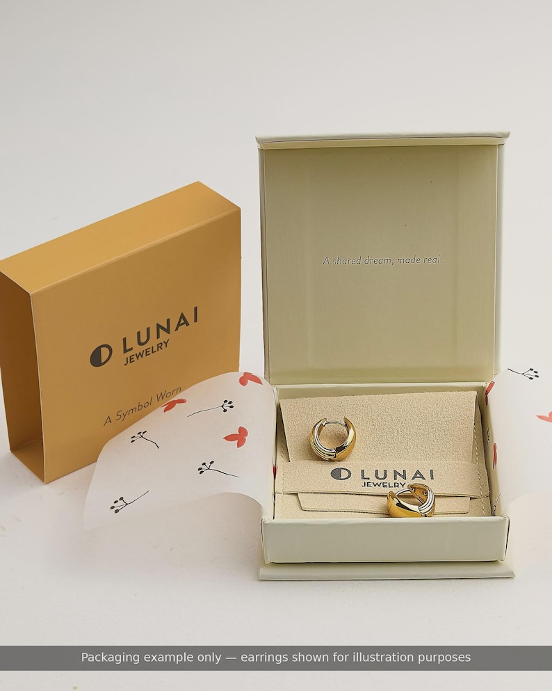 May include: A pair of two-tone gold and silver hoop earrings resting inside an open cream-colored jewelry box. The box lid features the text A shared dream, made real. The box includes a branded insert with the text LUNAI JEWELRY.