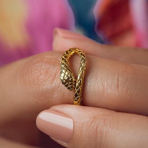 May include: A gold-coloured ring shaped like a coiled snake. The metal surface features a detailed, repeating scale pattern across the entire length of the band, with the snake's head clearly defined at one end of the open-style ring design.