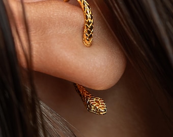 May include: A gold-toned ear cuff shaped like a slender snake that wraps around the ear cartilage. The jewelry features a detailed, textured scale pattern along its entire length, culminating in a defined snake head at the lower end.