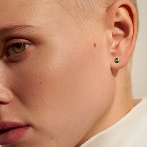May include: A small, circular stud earring featuring a vibrant blue gemstone set within a polished gold-toned metal bezel. The earring is worn in the earlobe, showcasing a simple, minimalist design that highlights the contrast between the gold and blue.