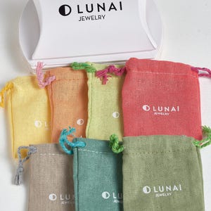 May include: A white pillow box with the black text LUNAI JEWELRY and a crescent moon icon, positioned above seven small fabric drawstring pouches in yellow, orange, lime green, coral, beige, teal, and olive green, each featuring the LUNAI JEWELRY logo.