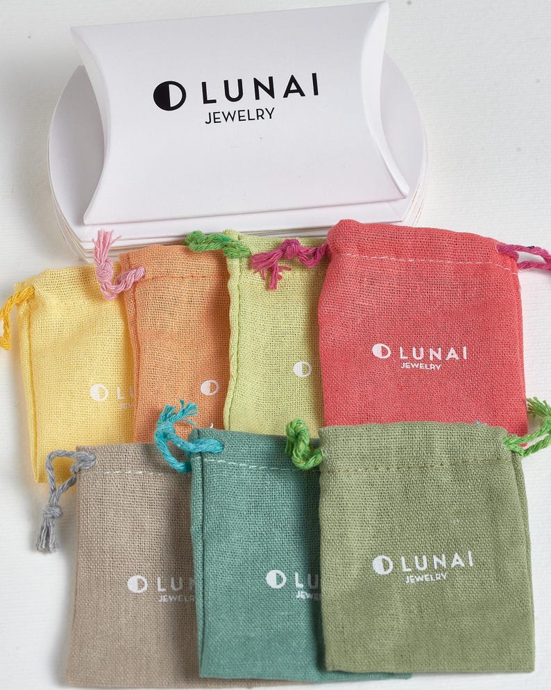 May include: A white pillow box with the black text LUNAI JEWELRY and a crescent moon icon, positioned above seven small fabric drawstring pouches in yellow, orange, lime green, coral, beige, teal, and olive green, each featuring the LUNAI JEWELRY logo.