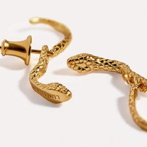 May include: A pair of gold-toned hoop earrings shaped like snakes with textured scales. One earring features a post and a bell-shaped back, while the other is shown from the side. The word LUNA is engraved on the underside of one snake head.