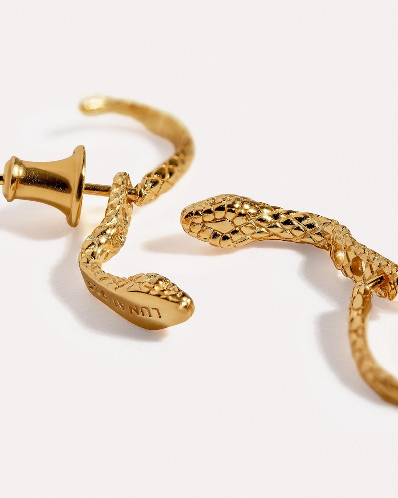 May include: A pair of gold-toned hoop earrings shaped like snakes with textured scales. One earring features a post and a bell-shaped back, while the other is shown from the side. The word LUNA is engraved on the underside of one snake head.