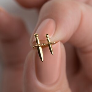 May include: A pair of gold-toned stud earrings shaped like miniature daggers. Each earring features a smooth, polished finish with a rounded pommel, a horizontal crossguard, and a pointed blade, creating a cohesive set of two small sword-like accessories.