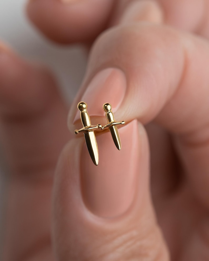May include: A pair of gold-toned stud earrings shaped like miniature daggers. Each earring features a smooth, polished finish with a rounded pommel, a horizontal crossguard, and a pointed blade, creating a cohesive set of two small sword-like accessories.