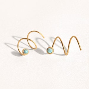 May include: Two gold-toned spiral earrings, each featuring a single round, iridescent light blue opal-like stone set at one end. The metal wire forms a continuous coil shape, creating a distinct, curved silhouette for each piece of jewellery.